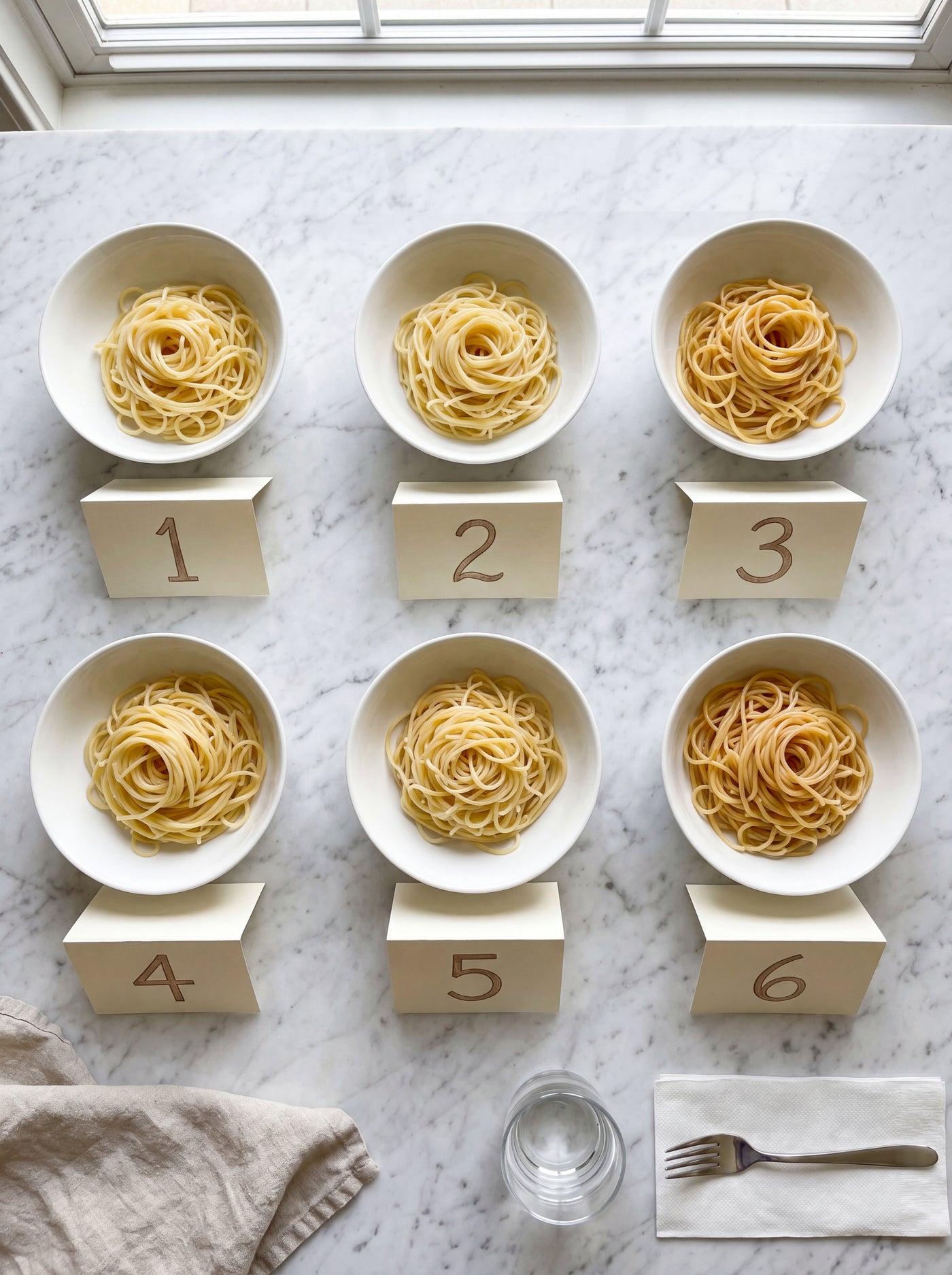 Overhead flat-lay of six numbered bowls of plain cooked spaghetti, the same pasta cut in every bowl, on a modern Carrara marble counter in a New York apartment for a blind tasting.