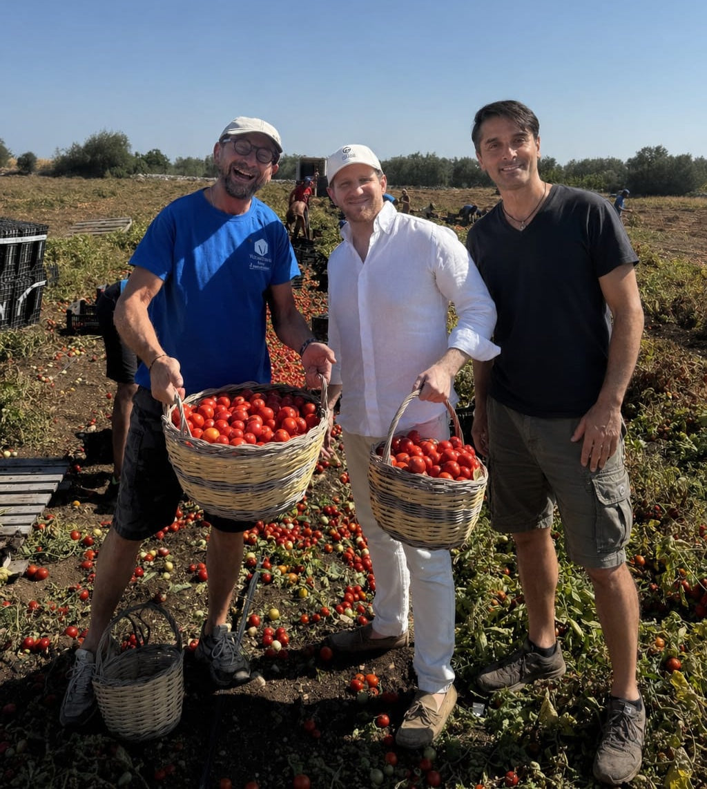 Sam and Alberto with a local grower in Puglia, holding baskets of just-harvested tomatoes in a sunlit field.